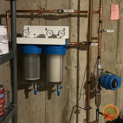 Home filtration A home water filtration system installed on a basement wall, featuring two large blue filter housings with pressure gauges mounted above each. Copper plumbing lines run vertically and horizontally, connecting to a water pump on the right. The setup is mounted on a concrete wall near a shelving unit with paint cans and other supplies. A circular logo is visible in the bottom right corner.